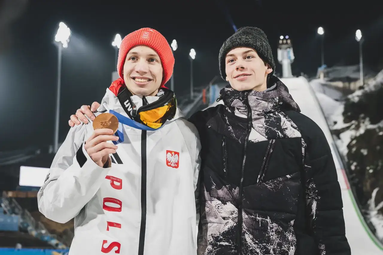 Kacper Tomasiak z rodziną świętuje medal. Dwóch młodych skoczków narciarskich, jeden z brązowym medalem, pozuje na tle skoczni.