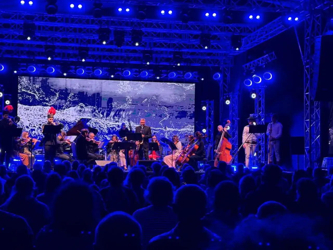 Koncert symfoniczny na festiwalu Kołobrzeg. Orkiestra gra na scenie oświetlonej niebieskimi światłami, a przed nią siedzi publiczność.