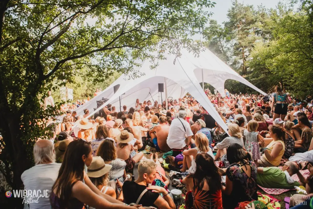 Tłum ludzi siedzi pod białym namiotem na festiwalu Wibracje, ciesząc się słońcem i muzyką.
