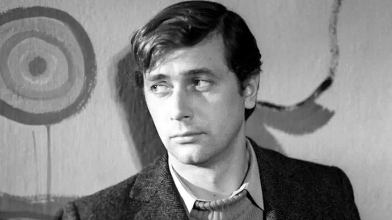 Młody Janusz Gajos, znany z wielu filmów, seriali i programów, spogląda w bok na tle abstrakcyjnego obrazu.