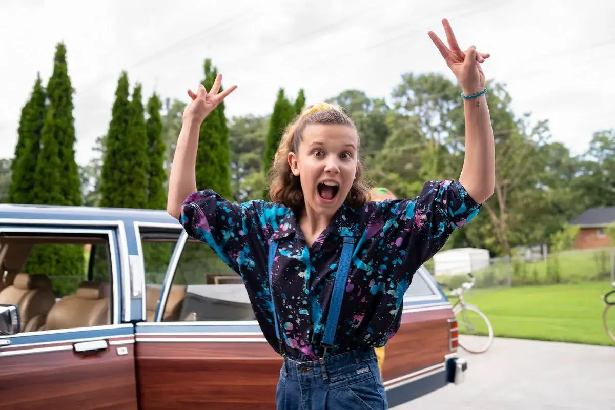 Nastka z Stranger Things w kolorowej koszuli i szelkach, z rękami w górze, radośnie pozuje obok starego samochodu.