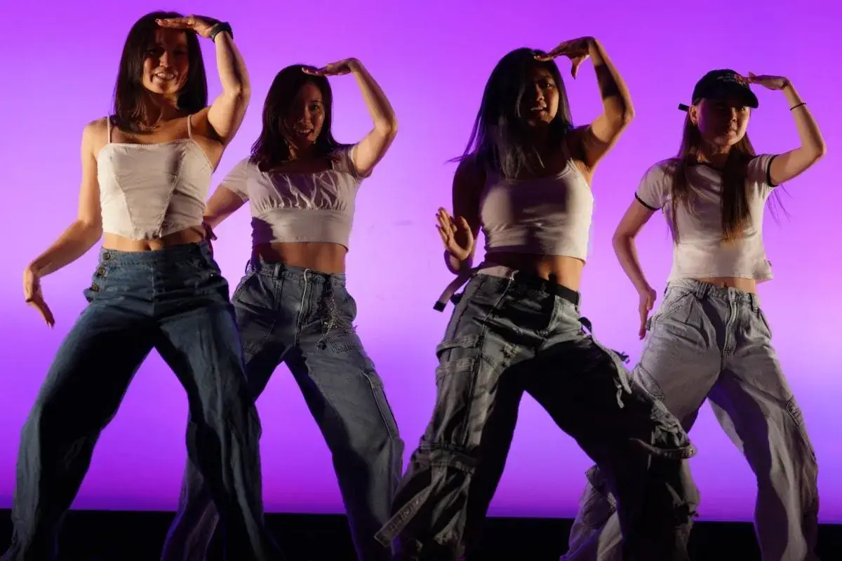 Cztery tancerki w białych topach i jeansach wykonują choreografię w stylu k-pop na fioletowym tle.