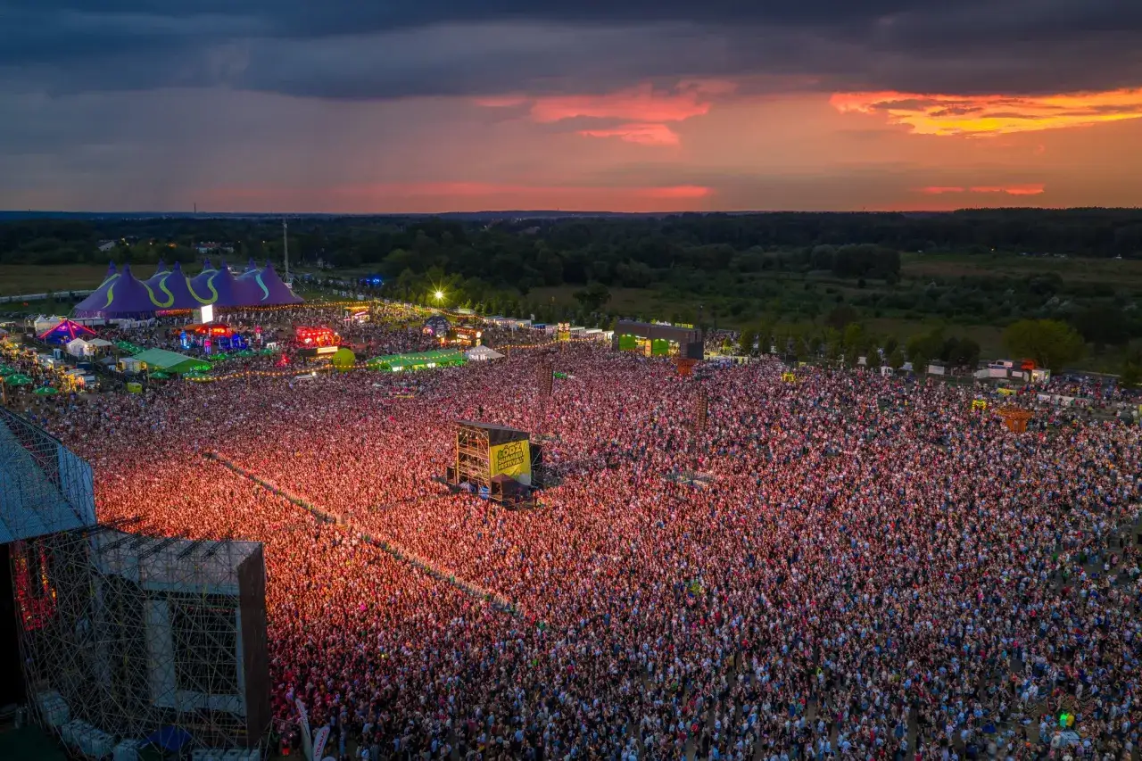 Tysiące ludzi na koncercie podczas Dni Łodzi. Zachodzące słońce maluje niebo na pomarańczowo i różowo nad tłumem zgromadzonym przed sceną.