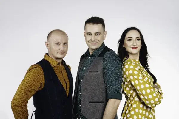 Trzy osoby: łysy mężczyzna w kamizelce, mężczyzna w zielonej koszuli i kamizelce, kobieta w żółtej sukience w grochy.