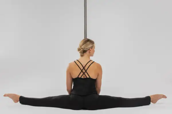 Kobieta w czarnym stroju siedzi w szpagacie przy rurze do pole dance.