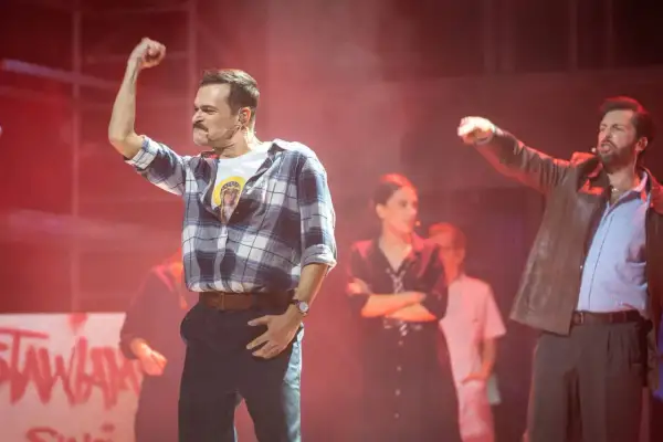 Aktorzy na scenie musicalu, jeden z podniesioną pięścią, w tle inne postacie i napisy.