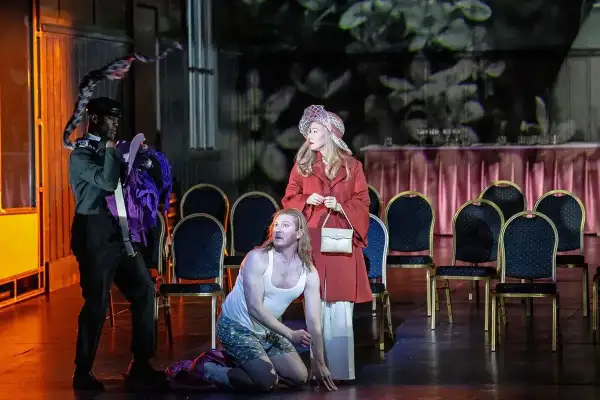 Aktorzy na scenie operetki, mężczyzna klęczy, kobieta stoi, trzeci mężczyzna trzyma kostium.
