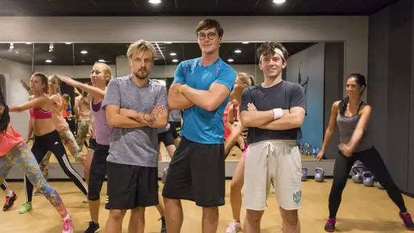 Trzech mężczyzn w strojach sportowych stoi z rękami skrzyżowanymi na piersi, w tle grupa kobiet ćwiczy.