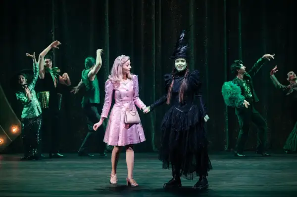 Elphaba w czarnej sukni i Glinda w różowej sukience trzymają się za ręce na scenie.