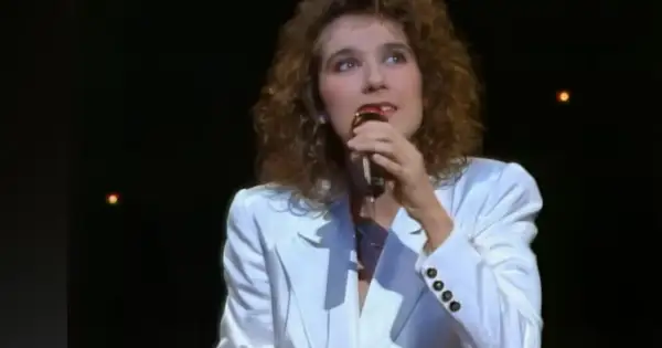Celine Dion w białej marynarce śpiewa do mikrofonu na ciemnej scenie.